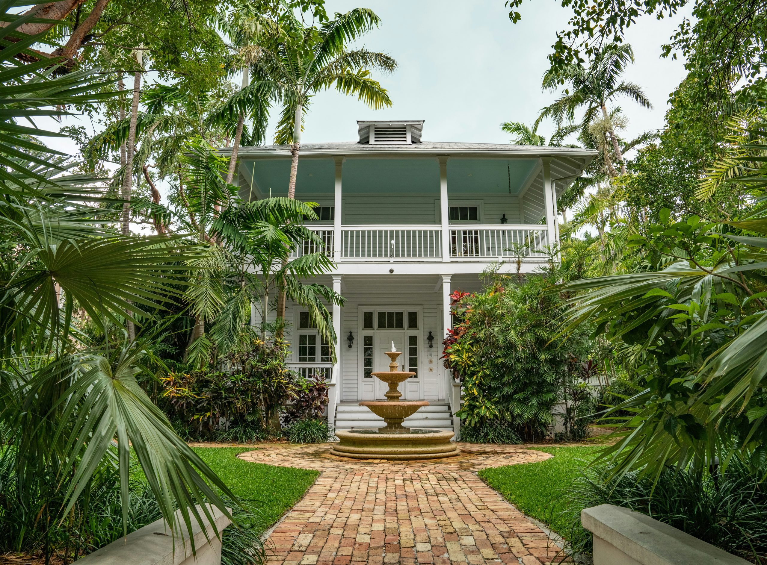Key West-style two-story home with white balcony, brick path, fountain and palm trees